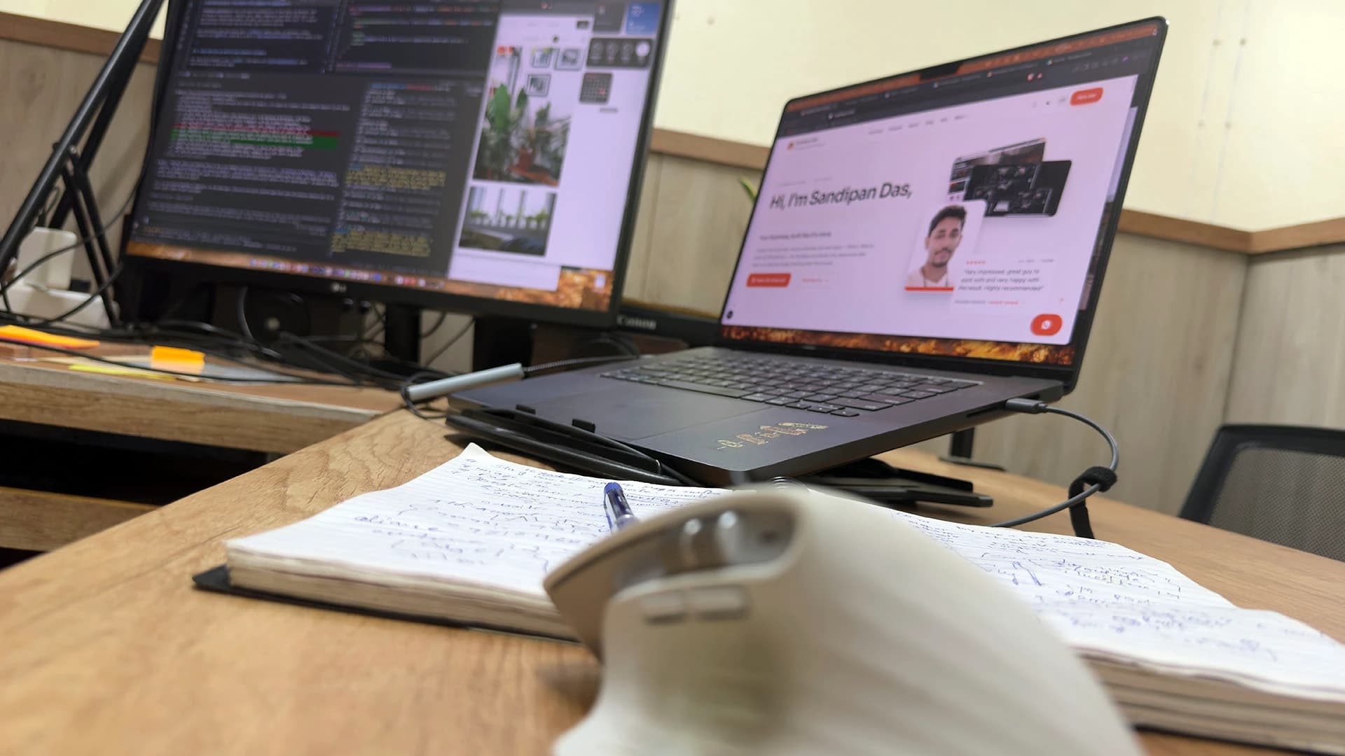 On a wooden desk in Kolkata, India: code on one screen, a website on a laptop, a notebook and a mouse in front